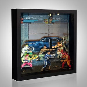Op de afbeelding: Een ingelijste 3D-schaduwbox met een retro videogame-scène met actiefiguren en een blauwe auto. De scène bevat de tekst "Punisher" en "Ka-Blam!" in een zwart frame.