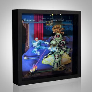 Puede incluir: Una obra de arte 3D enmarcada que representa una escena de videojuego. La obra muestra a un personaje con un traje azul y blanco pateando a un robot verde. El texto incluye "PUNISHER x1" y "NICK FURY x0". El marco es negro.