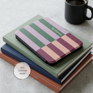 May include: A personalized tablet case with a geometric pattern in green, lavender, peach, and burgundy stripes. The name "CHARLOTTE" is printed on the case. The case rests on a stack of books, with a black mug of coffee in the background.