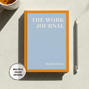 May include: A hardcover journal with a light blue cover and orange border. The text "THE WORK JOURNAL" is printed in white. The name "Madison Brown" is printed below. A gray pencil rests on the surface.