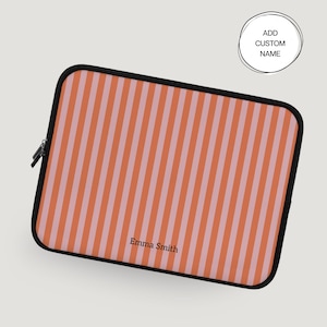 May include: A rectangular laptop sleeve with a vertical striped pattern in shades of orange and light pink. The sleeve has a black border and a zipper closure. The text "Emma Smith" is printed on the lower right corner. A circular graphic reads "Add Custom Name".