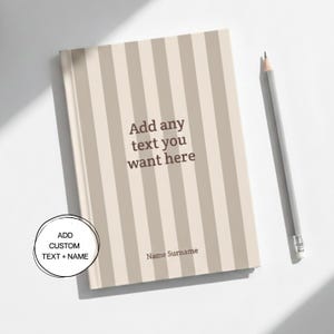 May include: A personalized notebook with vertical beige and off-white stripes. The cover features the text "Add any text you want here" and "Name Surname". A gray pencil rests beside the notebook. The notebook is designed for custom text and names.