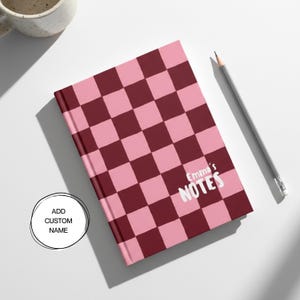 May include: A pink and burgundy checkered notebook with the text "Emma's NOTES" in white. A gray pencil rests beside the notebook. A white mug is in the upper left corner. The notebook cover has a checkerboard pattern.
