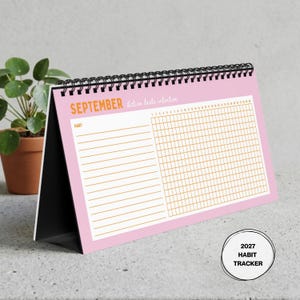 May include: A pink desk calendar with black spiral binding featuring the text SEPTEMBER and Action beats intention. The page includes a habit tracker grid with thirty-one columns and horizontal lines for habit names. A 2027 HABIT TRACKER label is visible.