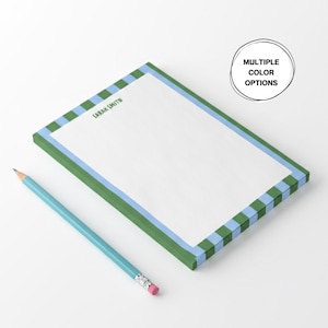 May include: A rectangular notepad with a green and blue striped border and the text "SARAH SMITH" in green. A light blue pencil with a pink eraser rests beside the notepad. The notepad has multiple color options.