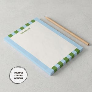 May include: A rectangular notepad with a white centre and a border featuring alternating light blue and green squares. The text MS MILLER is printed in green at the top. A wooden pencil lies diagonally next to the notepad. A circular badge reads MULTIPLE COLORS OPTIONS.