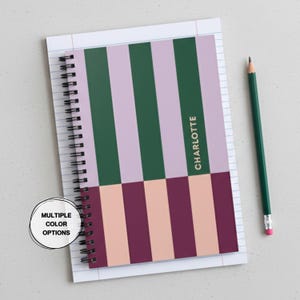 May include: A spiral-bound notebook with a colorful striped design in green, lavender, peach, and burgundy. The name "CHARLOTTE" is printed vertically on the right side. A green pencil with a pink eraser rests beside the notebook. The notebook is on lined paper.