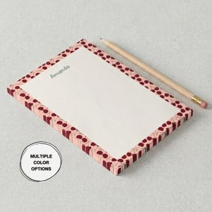 May include: A rectangular notepad with a pink border featuring a repeating pattern of maroon circles and shapes. The notepad has the name "Amanda" printed in green. A wooden pencil with a pink eraser rests on the side. The notepad is a stationery item.