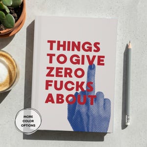 May include: A rectangular notebook with a light gray cover featuring the red text THINGS TO GIVE ZERO FUCKS ABOUT and a blue halftone graphic of a hand making a middle finger gesture. A gray pencil is positioned vertically to the right side.