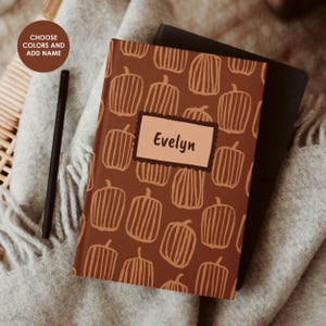 Puede incluir: Un cuaderno marrón con un patrón repetitivo de calabazas dibujadas a mano en color marrón claro. Una etiqueta rectangular en el centro muestra el nombre Evelyn en texto marrón oscuro. Un bolígrafo negro se sitúa a la izquierda del cuaderno.