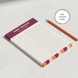 May include: A habit tracker notepad with the text "HABIT TRACKER SARAH BROWN". The notepad has a burgundy top and a color block design on the bottom edge. An orange pencil rests beside the notepad. The notepad is designed for tracking habits.