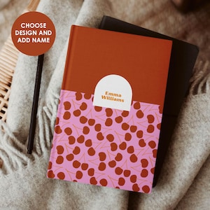 Op de afbeelding: Een gepersonaliseerd notitieboek met een kersenpatroon op een roze achtergrond. Het bovenste gedeelte is effen oranjerood van kleur. De tekst "Emma Williams" staat op een wit label. Het notitieboek ligt op een zachte, neutraal gekleurde deken.