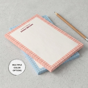 May include: Two rectangular notepads, one pink and one blue, with a grid pattern border. The top notepad has the text "A note from Emma Brown." A pencil rests on the surface. The image also includes a "Multiple Color Options" graphic.