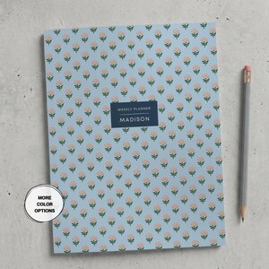 May include: A light blue weekly planner with a repeating pattern of small pink flowers and green stems. A dark blue rectangular label in the center features the text "WEEKLY PLANNER" and "MADISON" in white. A gray pencil is positioned to the right.