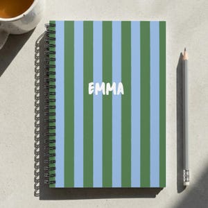 May include: A spiral-bound notebook featuring a vertical striped pattern in light blue and dark green. The name EMMA is printed in white capital letters across the center of the cover. A gray pencil is positioned to the right of the notebook.