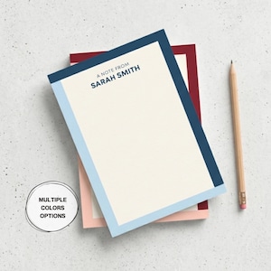 Puede incluir: Una pila de blocs de notas con interior color crema y borde azul, celeste y burdeos. El bloc de notas superior tiene el texto "A note from Sarah Smith" impreso en azul marino. Un lápiz de madera con goma de borrar rosa descansa al lado.