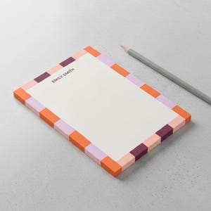 May include: A rectangular notepad with a white center and a border featuring a repeating pattern of orange, light pink, lavender, and dark purple blocks. The name EMILY SMITH is printed in dark purple text at the top. A gray pencil lies nearby.