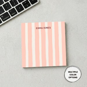 May include: A square notepad with a pink and white striped pattern. The text "EMMA JONES" is printed in a dark red font at the top. A keyboard is visible in the background. The notepad is on a gray surface. The notepad has multiple color options.