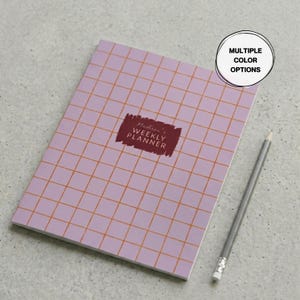 May include: A lavender weekly planner with an orange grid pattern and the text "Madison's Weekly Planner" in a burgundy rectangle. A gray pencil rests beside the planner. The image also includes a circle with the text "MULTIPLE COLOR OPTIONS".