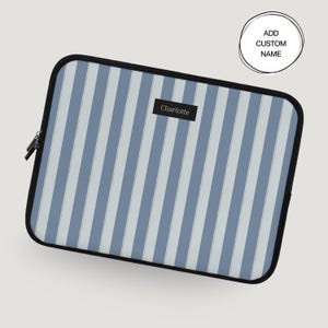 May include: A laptop sleeve with a black zipper and trim. The sleeve features a vertical striped pattern in light and medium blue. A small black rectangle with the name "Charlotte" is centered on the sleeve. A circular graphic reads "Add Custom Name."