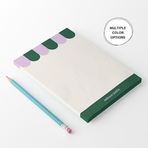 Puede incluir: Un bloc de notas rectangular con un diseño festoneado verde y lila en el borde superior. El borde inferior es verde con el nombre "MIRIAM SMITH" impreso en blanco. Un lápiz azul claro con una goma rosa descansa sobre el bloc de notas. El texto "MULTIPLE COLOR OPTIONS" está en un círculo.