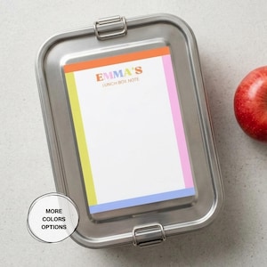 May include: A rectangular stainless steel lunch box with a metal latch closure. A white note card is attached to the lid, featuring a colorful border in orange, yellow, pink, and blue, with the text EMMA'S LUNCH BOX NOTE printed at the top.