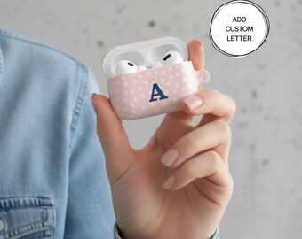 Puede incluir: Funda para AirPod rosa con lunares y una letra "A" azul marino. La funda está abierta, revelando auriculares blancos en el interior. El texto "ADD CUSTOM LETTER" está en un círculo en el fondo.