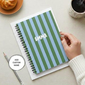May include: A spiral-bound notebook featuring a vertical striped pattern in alternating shades of light blue and dark green. The name EMMA is printed in white capital letters across the center of the cover. A gray pencil lies nearby.