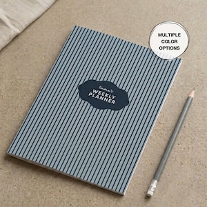 May include: A weekly planner with a blue and white striped pattern. The planner has the text "Emma's Weekly Planner" on a dark blue cloud-shaped label. A gray pencil rests on the surface next to the planner. The image also includes a "Multiple Color Options" label.