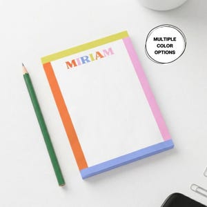 Pode incluir: Um bloco de notas personalizado com o nome "MIRIAM" em letras coloridas. O bloco de notas tem um interior branco com uma parte superior amarela, um lado esquerdo laranja, um lado direito rosa e uma parte inferior azul. Um lápis verde repousa sobre a superfície branca.
