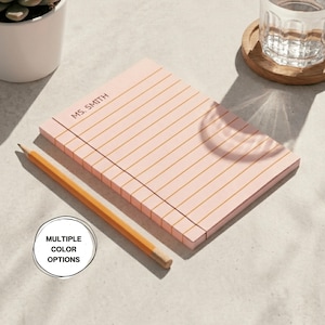 May include: A pink notepad with gold horizontal lines and the text "MS. SMITH" printed at the top. A yellow pencil rests on the notepad. The image also includes a glass of water and a plant.