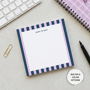 May include: A square notepad with a white interior and the text "NOTE TO SELF". The notepad has a navy blue border with pink and navy blue stripes. A gold paperclip, a black pen, a keyboard, and a spiral notebook are also visible.