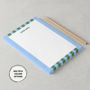 May include: A rectangular notepad with a white center and a border featuring alternating light blue and green stripes. The name Sarah Smith is printed in green at the top. A wooden pencil lies diagonally next to the notepad. A circular graphic contains the text Multiple Colors Options.