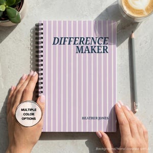 Könnte beinhalten: Ein spiralgebundenes Notizbuch mit einem hellviolett-weiß gestreiften Einband. Der Text "DIFFERENCE MAKER" ist oben in Dunkelblau gedruckt, und der Name "HEATHER JONES" steht unten rechts ebenfalls in Dunkelblau.