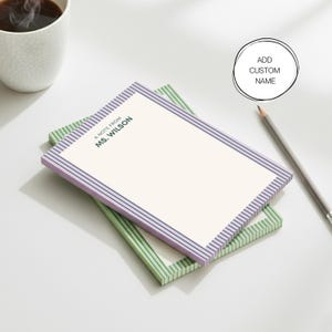 Op de afbeelding: Twee gepersonaliseerde notitieblokken, één lavendelkleurig en één groen, met gestreepte randen. Op het bovenste notitieblok staat de tekst "A note from Ms. Wilson" in donkergroen. Een potlood en een kopje koffie zijn ook zichtbaar.