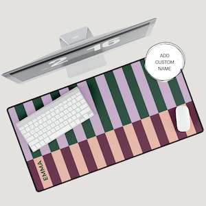 May include: A rectangular desk mat with a colorful striped pattern in green, lavender, and peach. A white keyboard and mouse are on the mat, with the name "EMMA" printed on the bottom left. A computer monitor is above the mat, and a circle with the text "ADD CUSTOM NAME" is on the right.
