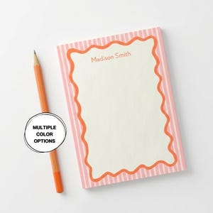 Może przedstawiać: Prostokątny notatnik z różowo-białą obwódką w paski i falistą pomarańczową obwódką. Na notatniku u góry wydrukowano imię i nazwisko "Madison Smith". Obok notatnika umieszczono pomarańczowy ołówek. Tekst "MULTIPLE COLOR OPTIONS" znajduje się w okręgu.