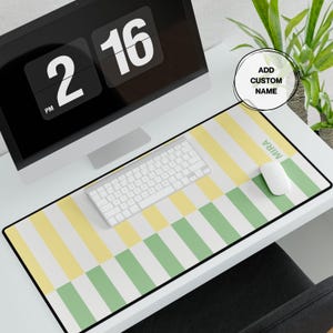 May include: A rectangular desk mat featuring a repeating vertical striped pattern in pale yellow, sage green, and white. The mat includes the custom name MIRA printed in a light green sans-serif font on the right side, with a black border edge.