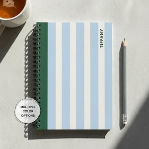 May include: A spiral-bound notebook featuring vertical stripes in light blue, white, and a dark green edge. The name TIFFANY is printed in black on the cover. A circular badge states MULTIPLE COLOR OPTIONS. A gray pencil lies to the right.