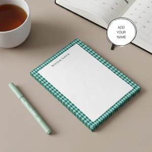 Puede incluir: Un bloc de notas personalizado con un borde a cuadros verdes y blancos. El bloc de notas tiene el nombre "Miriam Smith" impreso en la parte superior. También se ve un bolígrafo verde y una taza de té.