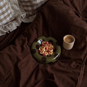 Puede incluir: Un cuenco de cerámica verde oliva con lunares de color marrón oscuro que contiene rodajas de plátano, frambuesas y nueces. Junto al cuenco hay una pequeña taza de cerámica de color crema con café, sobre una superficie de tela marrón oscuro.
