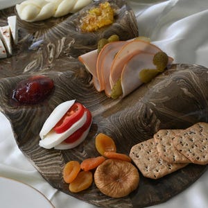 Puede incluir: Una bandeja de servir de madera con un patrón de mármol, llena de varios bocadillos, incluyendo pavo en rodajas, queso, galletas, fruta seca y mermelada.