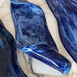 Könnte beinhalten: Drei handgefertigte Servierschalen in abstrakter Form und tiefblauen Farbtönen. Die Keramikstücke haben eine glänzende Oberfläche mit Marmoreffekt, was einen einzigartigen Look erzeugt. Diese dekorativen Schalen eignen sich ideal zum Servieren von Vorspeisen oder zum Präsentieren kleiner Gegenstände.