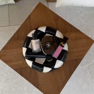 Könnte beinhalten: Eine runde Keramikschale mit schwarz-weißem Schachbrettmuster, die verschiedene Kosmetikartikel enthält, darunter ein Chanel-Puder, eine Foundation-Flasche, ein rosa Lipgloss und eine kleine schwarze Tube, die in der Mitte angeordnet sind.