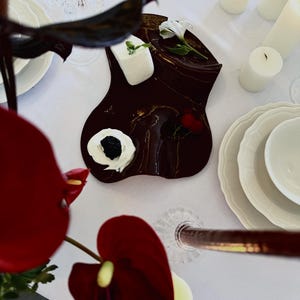 Pode incluir: Uma bandeja de servir castanho-escura de forma orgânica contém um bloco retangular de queijo branco, uma porção de natas batidas com uma amora, um pequeno cacho de cerejas vermelhas e uma flor branca sobre uma toalha de mesa branca.