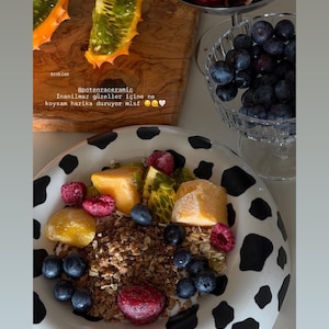 May include: A round ceramic bowl with a black and white cow print pattern containing a mixture of granola, fresh blueberries, raspberries, and cubed yellow fruit. The bowl is positioned below a wooden board with sliced kiwano fruit and glass bowls.