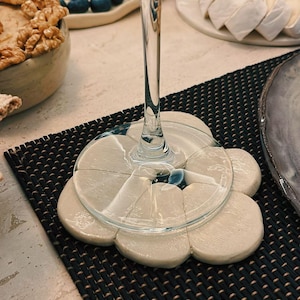 May include: A stemmed wine glass sits on a white, flower-shaped coaster. The coaster is made of several pieces and rests on a black woven placemat. The background includes a plate of cheese and blueberries.