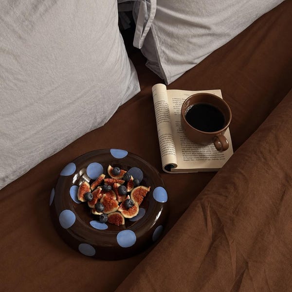 May include: A dark brown ceramic bowl with light blue polka dots containing sliced figs and blueberries, positioned next to an open book and a dark brown mug filled with black coffee on a dark brown fabric surface with light gray pillows.