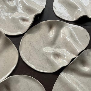 Può includere: Una collezione di piatti in ceramica fatti a mano, di forma organica. I piatti sono di colore bianco sporco macchiettato con finitura lucida. Ogni piatto ha una forma ondulata unica, che suggerisce un design naturale e fluido. I piatti sono disposti su una superficie scura.