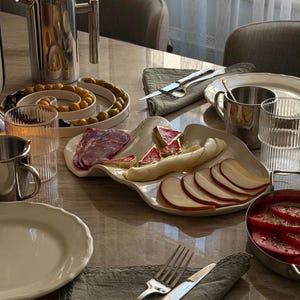 Könnte beinhalten: Ein gedeckter Tisch mit einer French Press aus Edelstahl, einer weißen Spiralschale mit grünen und schwarzen Oliven, einer weißen gewellten Platte mit Apfelscheiben, Käseecken und Aufschnitt sowie Tellern, Gläsern und Besteck.